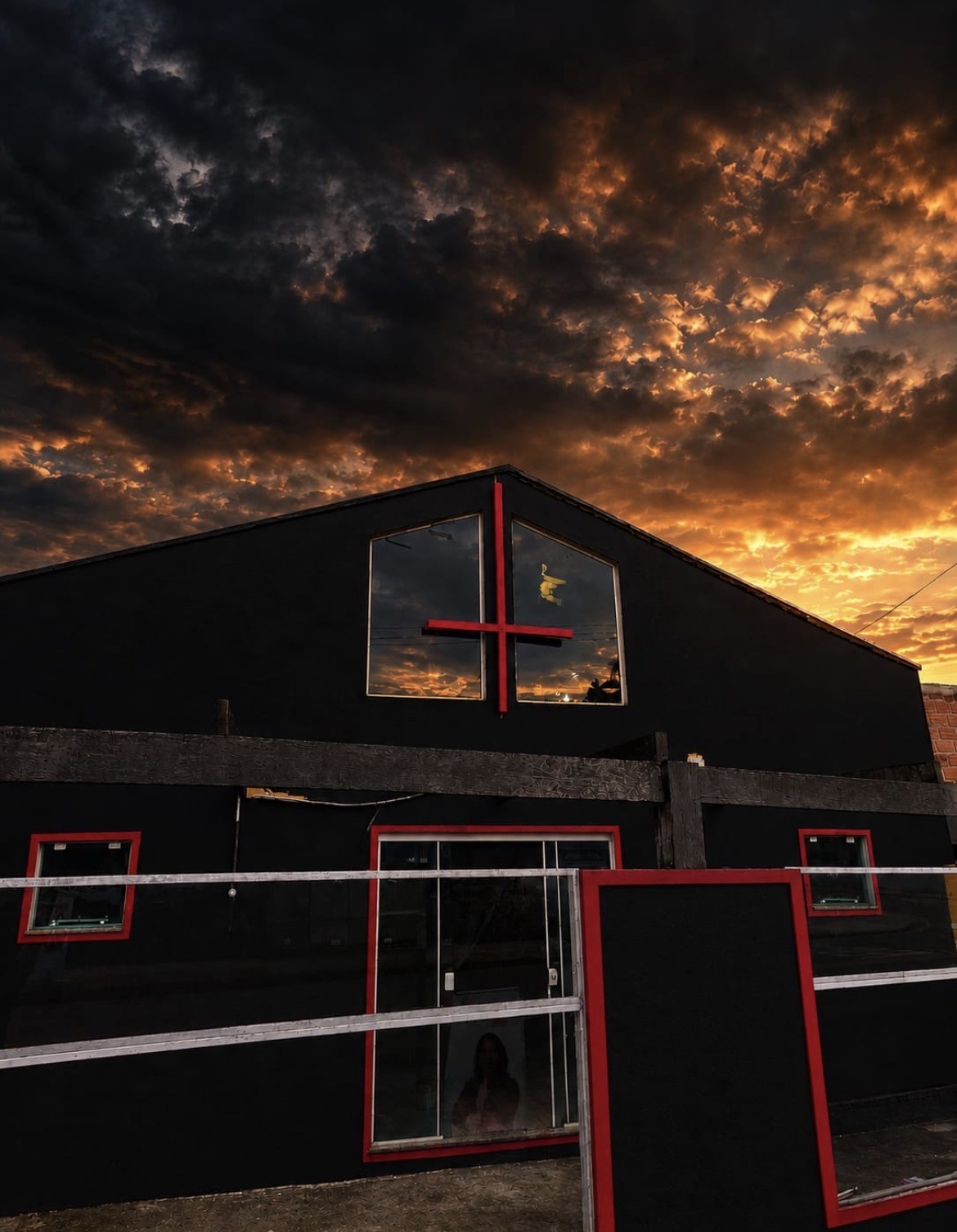 Fachada da Igreja de Lúcifer Curitiba, em preto e vermelho, com uma cruz vermelha e céu iluminado ao fundo